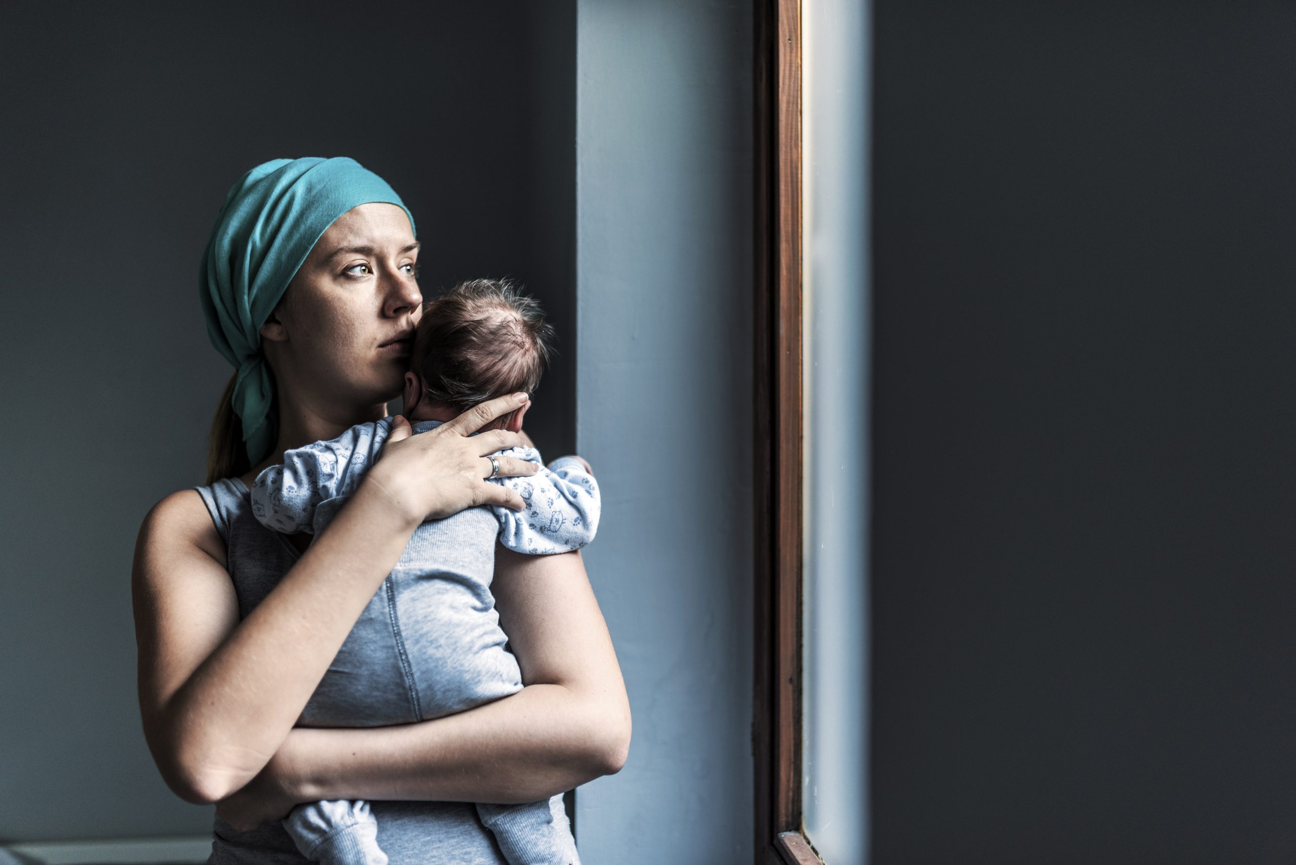 Caucasian Pensive Woman in headscarf, fighting breast cancer while holding her newborn baby relaxing in cancer treatment hospital, patient standing next to hospital window. Mother and baby son. Sleepy little child with serious mom.